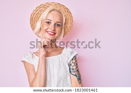 Young blonde woman with tattoo wearing summer hat smiling looking confident at the camera with crossed arms and hand on chin. thinking positive. 