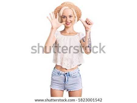 Young blonde woman with tattoo wearing summer hat showing and pointing up with fingers number six while smiling confident and happy. 