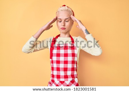 Young blonde woman with tattoo wearing professional baker apron with hand on head, headache because stress. suffering migraine. 