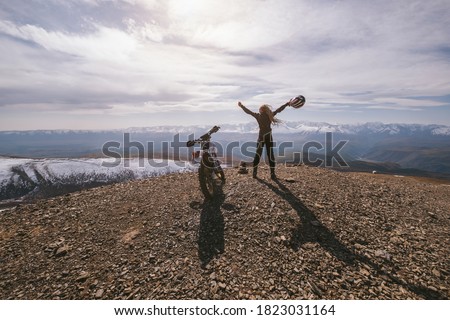 Female motorcyclist standing with her enduro motorcycle on mountain top, snow peaks skyline view