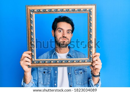 Young hispanic man holding empty frame puffing cheeks with funny face. mouth inflated with air, catching air. 