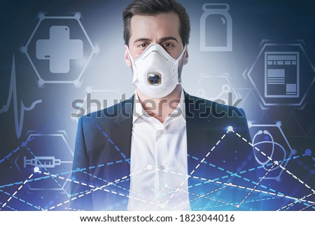 Young European businessman standing in protective mask over blurry blue background with double exposure of medical icons and graphs. Toned image