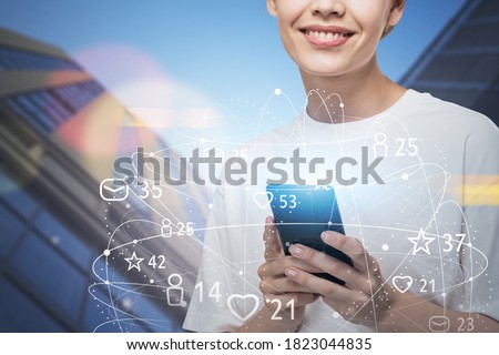 Cheerful young woman with smartphone standing in city with double exposure of social media icons. Concept of job search. Toned image