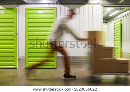 Blurred motion shot of unrecognizable man pushing cart with boxes while running in self storage facility, copy space Royalty-Free Stock Photo #1823056022