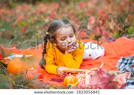 A little pretty girl sits in the park on an orange rug and looks at a pumpkin.
