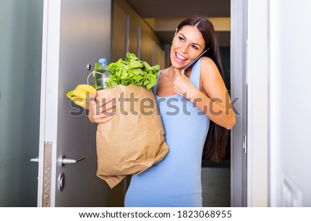 Pretty woman arriving home carrying groceries in a paper bag opening the door and on a phone call - Multi tasking