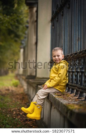 The boy sits by the fence in the park. The boy is wearing yellow clothes. Autumn photo session.