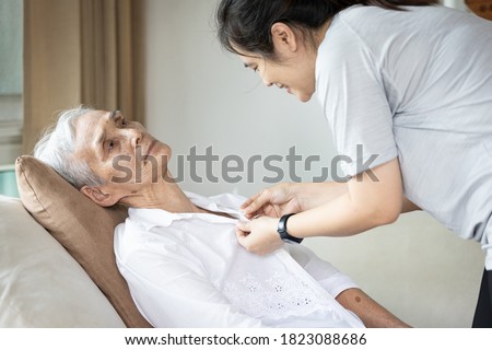Asian female caregiver taking care of helping elderly patient get dressed,button on the shirt or changing clothes for a paralyzed person,senior woman with paralysis of limbs,body or muscles weakness Royalty-Free Stock Photo #1823088686