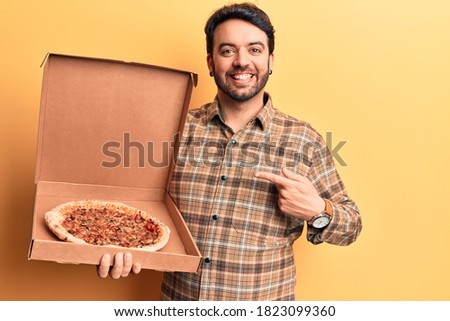 Young hispanic man holding delivery pizza box smiling happy pointing with hand and finger 