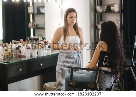 Beautiful female makeup artist doing makeup for a young brunette hair girl in a beauty salon sitting in front of a large mirror. Concept of preparation for the holiday and meeting. Start working