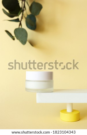 Mockup moisturizer cream in jar standing on abstract pedestal on pastel yellow background with copy space and defocused eucalyptus leaves, front view. Skincare beauty product template, vertical