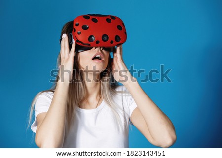 Young woman holds virtual reality glasses in her hands scared and screaming. Computer games, blue background, emotions.