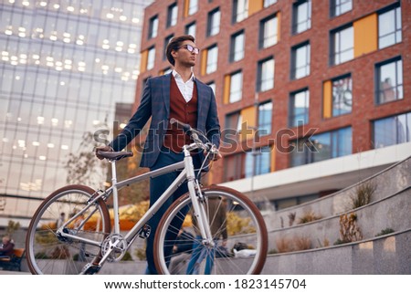 Low angle of young businessman in smart casual clothes standing near bike on city street