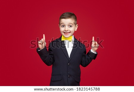 Happy boy in jacket and bow tie smiling from camera and pointing up against red background