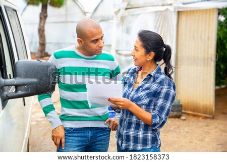 Latino man and woman farmers signing papers and communicating near car at glasshouse