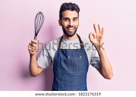 Young handsome man with beard wearing apron holding whisk doing ok sign with fingers, smiling friendly gesturing excellent symbol 