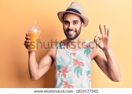 Young handsome man with beard wearing summer hat and t-shirt drinking orange juice doing ok sign with fingers, smiling friendly gesturing excellent symbol 