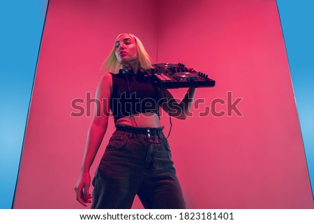 Young female musician in headphones performing on blue-red background in neon light. Concept of music, hobby, festival, entertainment, emotions. Joyful party host, DJ, portrait of artist.