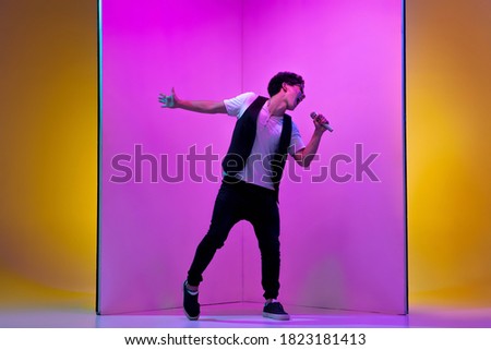 Young male musician, singer performing on pink-orange background in neon light. Concept of music, hobby, festival, entertainment, emotions. Joyful party host, singer, portrait of artist.