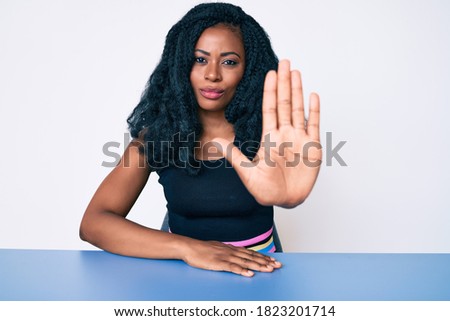 Beautiful african woman wearing casual clothes sitting on the table doing stop sing with palm of the hand. warning expression with negative and serious gesture on the face. 