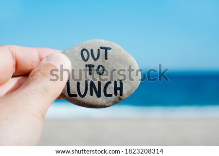 closeup of the hand of a young caucasian man on the beach, in front of the ocean, holding a stone with the text out to lunch written in it