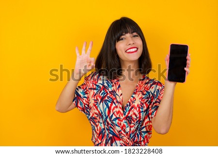 Excited woman showing smartohone blank screen, blinking eye and doing ok sign with hand. Studio shot of shocked girl holding smartphone with blank screen. Advertisement concept. Royalty-Free Stock Photo #1823228408
