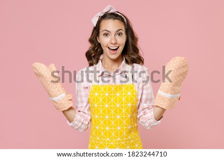 Surprised excited cheerful young brunette woman housewife 20s wearing yellow apron gloves potholders doing housework isolated on pastel pink colour background studio portrait. Housekeeping concept Royalty-Free Stock Photo #1823244710