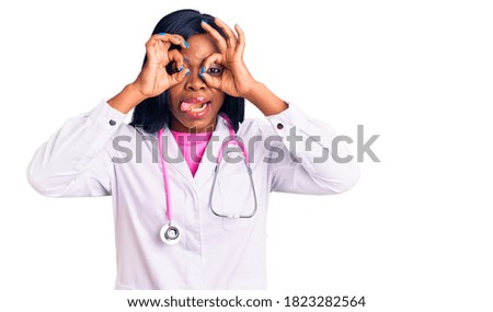 Young african american woman wearing doctor stethoscope doing ok gesture like binoculars sticking tongue out, eyes looking through fingers. crazy expression. 