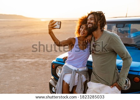 Image of joyful african american couple taking selfie on cellphone while travelling with car in desert