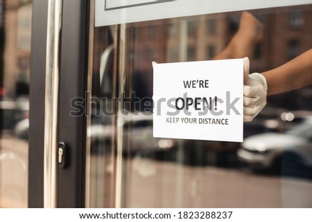 Social distancing, reopening and return to work after lockdown. Hands of african american woman in gloves hang paper with inscription, we are open, keep your distance, close up, free space, cropped