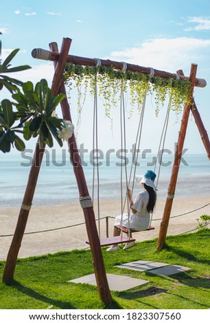 Picture from the back of a woman sitting on a swing that is tied to the bar in the morning. The top has ivy and the lower has green grass. Blurred backdrop is a blue ocean with glitter and clear sky.