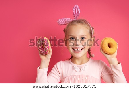 Portrait of a cheerful little girl with a funny headband having fun towards the camera with colorful donuts isolated on a pink background. Expression of true positive emotions of a cute child.