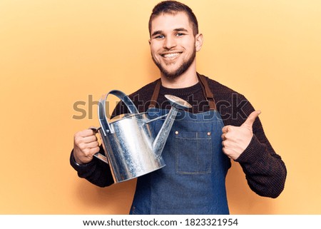 Young handsome man wearing gardener apron holding watering can smiling happy and positive, thumb up doing excellent and approval sign 