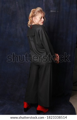 Fashion model dressed in grey loose comfortable suit decorated with brooches posing in studio with blue fabric background. Examples of autumn female fashion. Copy space. Full length back side view