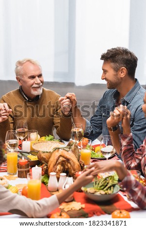 pleased multicultural family holding hands during dinner on thanksgiving holiday