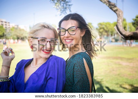 a young blonde woman in blue dress and a brunette woman in dark green dress pose in a selfie in a close up portrait with eyeglasses in a city park on a sunny day