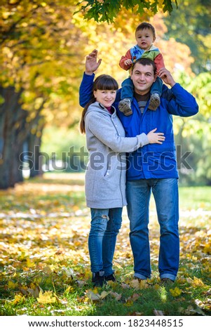 Family of three walks in the autumn park holding hands. happy father carrying son with maple leaves. Mother embrace her cute boy child. Happy family leisure together concept.