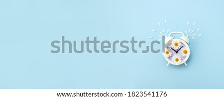 Alarm clock decorated with flowers and petals of chamomile on a light blue background. New day start banner. Copy space. Selective focus