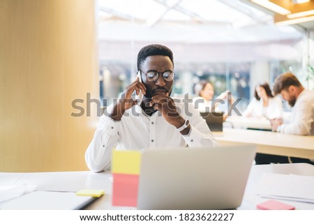Black formal man in eyeglasses hand talking on phone and contemplating on problem while sitting in front of laptop in office