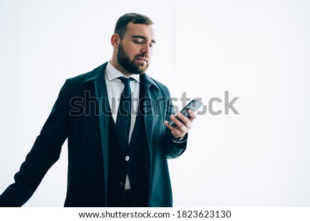 Formally dressed male entrepreneur using cellular application for checking account balance near publicity area, Caucasian businessman messaging via smartphone near mockup backdrop with copy space area