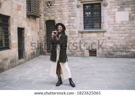 Full body of happy black female traveler in trendy wear and hat standing on street and taking picture on photo camera during stroll in Barcelona