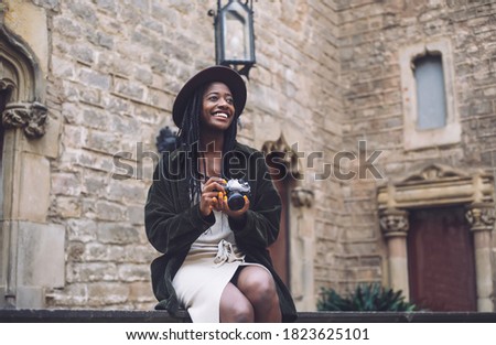 Positive young woman in trendy outfit holding old photo camera and looking away with toothy smile while sitting in yard of old historical building in Barcelona