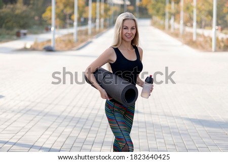 A beautiful blonde goes to a workout in the park outdoors and carries a rolled up yoga mat with her.