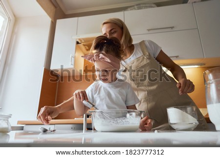 happy mother baking with little daughter in apron and cook hat working with flour , bowl and spoon preparing dough teaching the kid baking and having fun together.