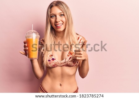 Young beautiful blonde woman wearing bikini drinking orange juice smiling happy and positive, thumb up doing excellent and approval sign 