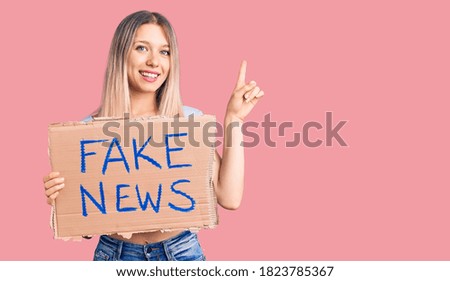 Young beautiful blonde woman holding fake news banner surprised with an idea or question pointing finger with happy face, number one 