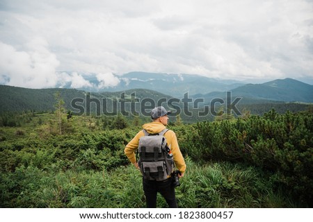 Young stylish guy photographer with a backpack and a camera stands enjoying and is inspired by the amazing mountain landscape