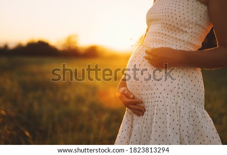 Close-up of pregnant woman with hands on her belly on nature background. Silhouette of pregnant woman in white dress in sunlight of sunset. Concept of pregnancy, maternity, expectation for baby birth.