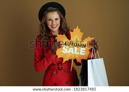Hello october. happy elegant middle aged housewife in red coat and black beret with autumn sale leaf shape banner, leather gloves and paper shopping bags isolated on beige background.