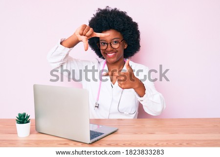Young african american woman wearing doctor stethoscope working using computer laptop smiling making frame with hands and fingers with happy face. creativity and photography concept. 